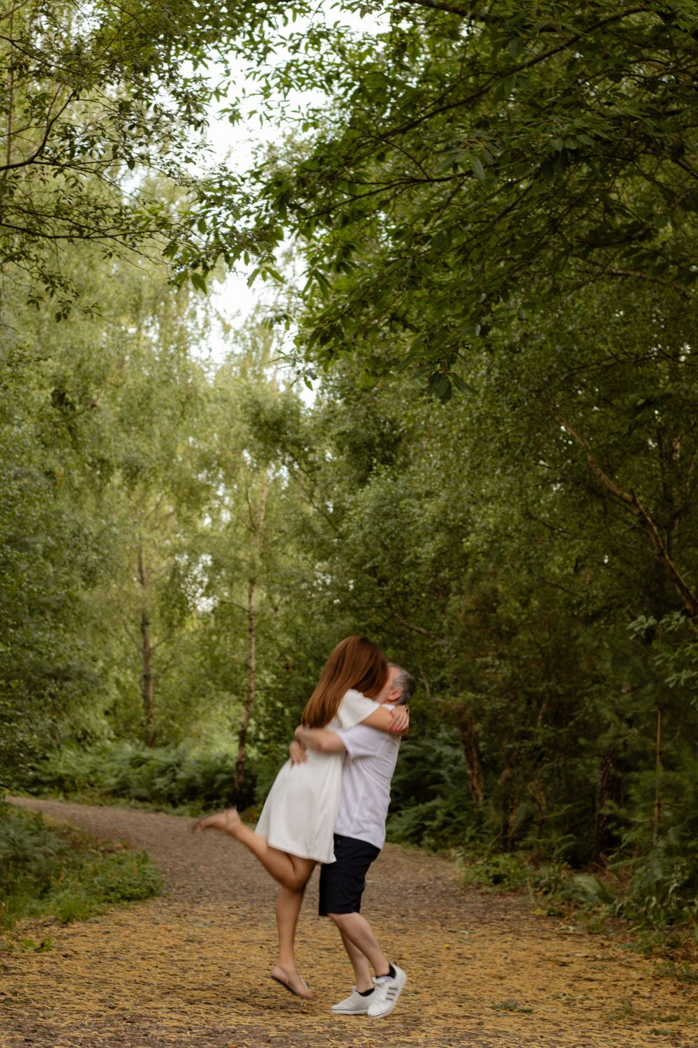 Couple embracing and hugging on a forest path surrounded by lush green trees in a romantic outdoor setting.