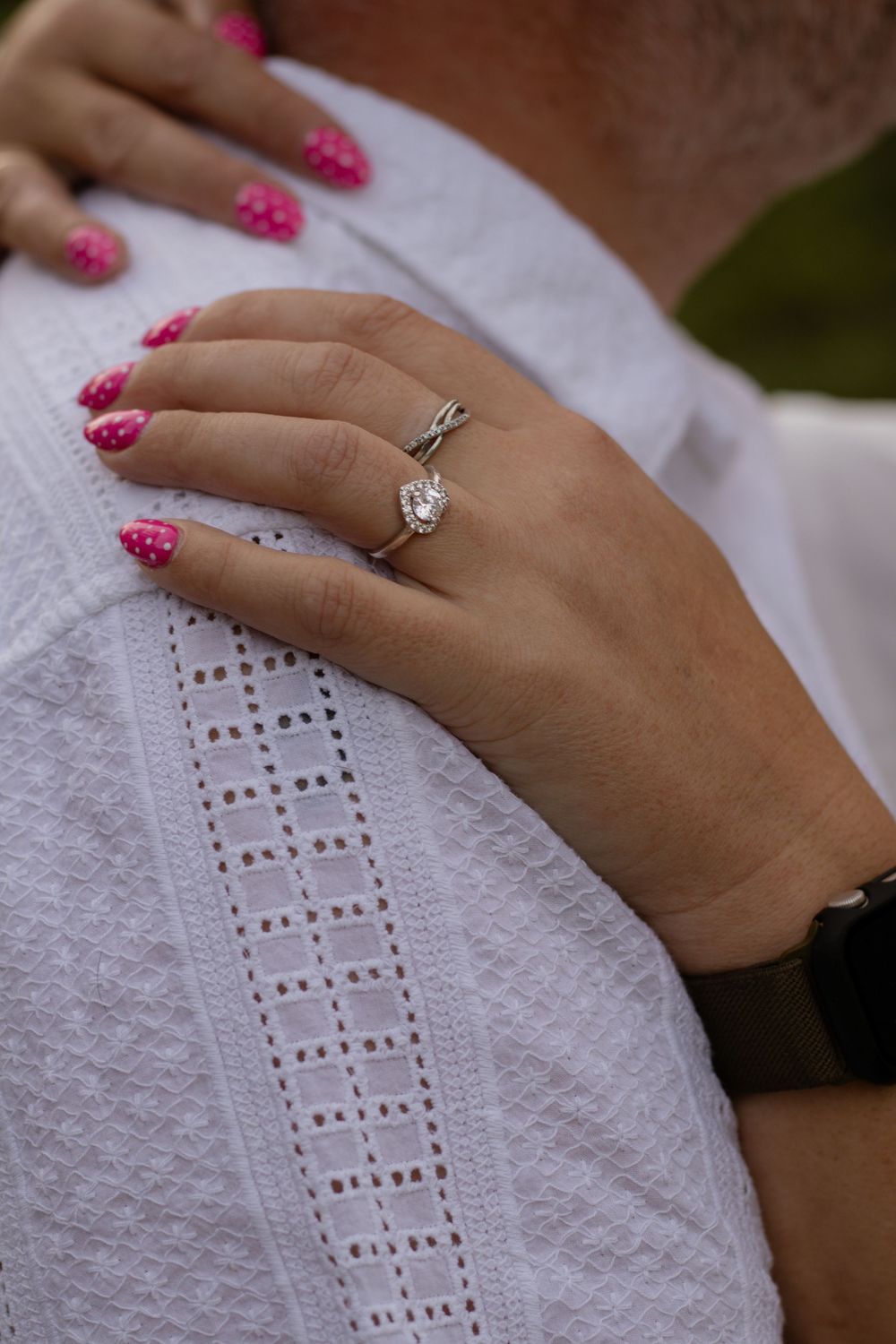 Close-up of a woman's hand with pink nails wearing a diamond engagement ring, resting on a man's white shirt.