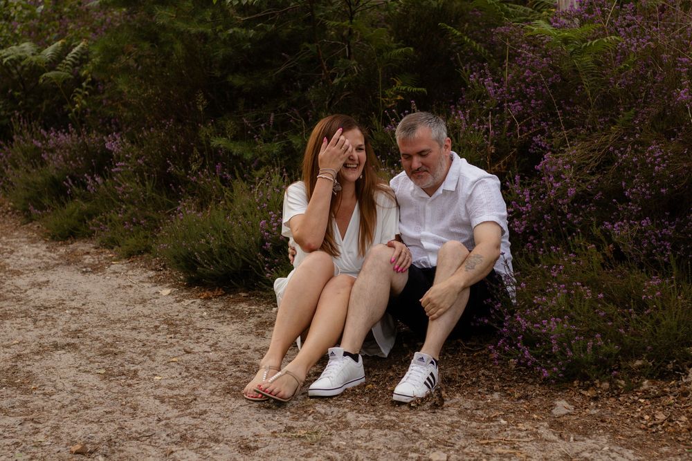 A couple sits together on a sandy path surrounded by purple heather, sharing a joyful moment in nature.