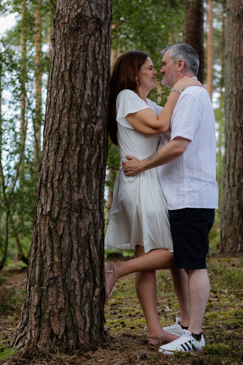 A romantic couple embracing and smiling by a pine tree in a forest, woman in white dress, man in white shirt.