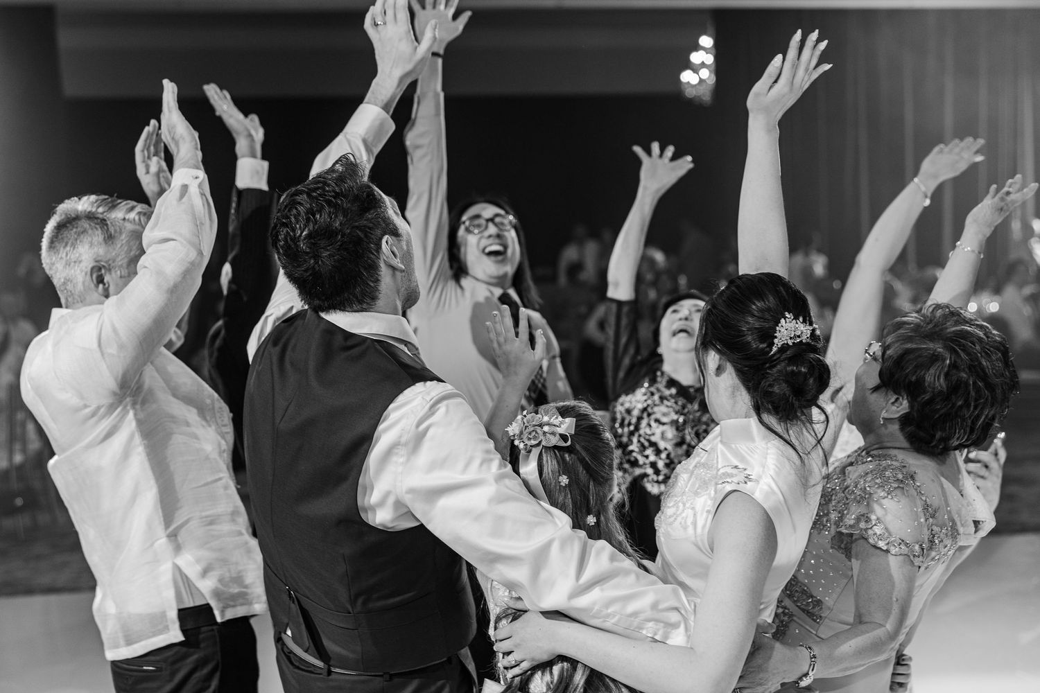 Black and white photo of joyful wedding guests dancing with arms raised at a lively wedding reception celebration.