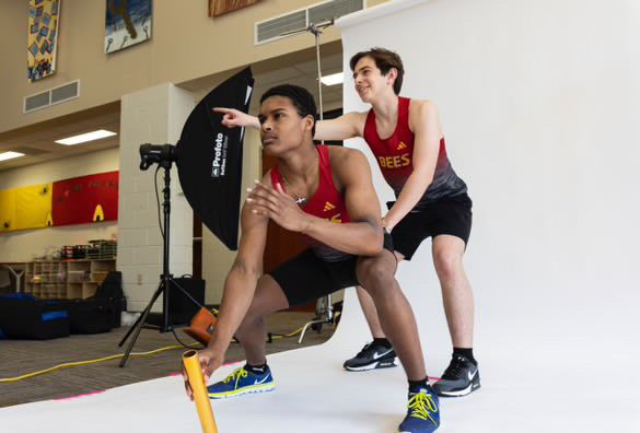 Two athletes in red uniforms practice sprinting form indoors near photography equipment and a starting block.