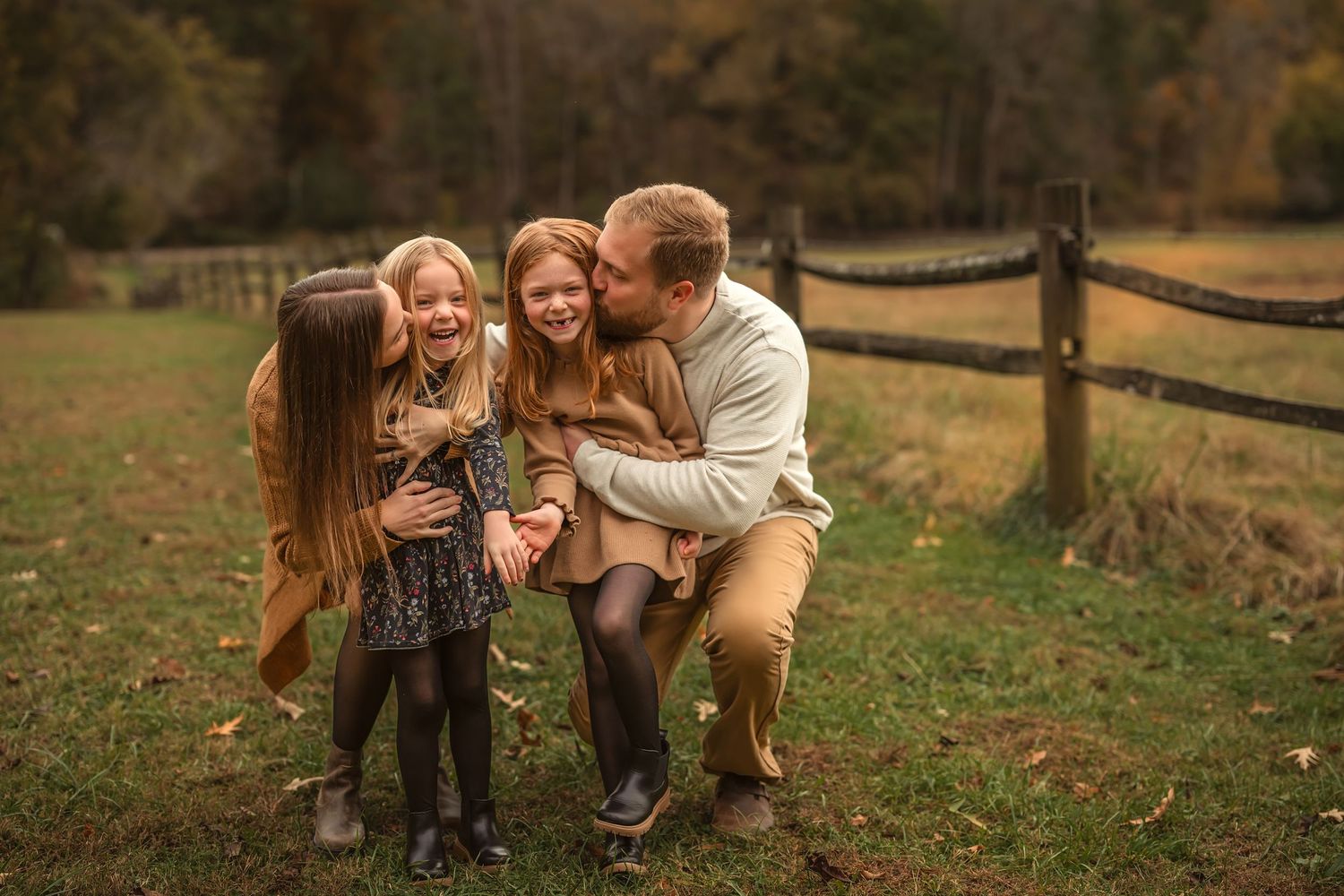Family of four laughing together outdoors in autumn, father crouching with two daughters and mother in a rustic field setting.