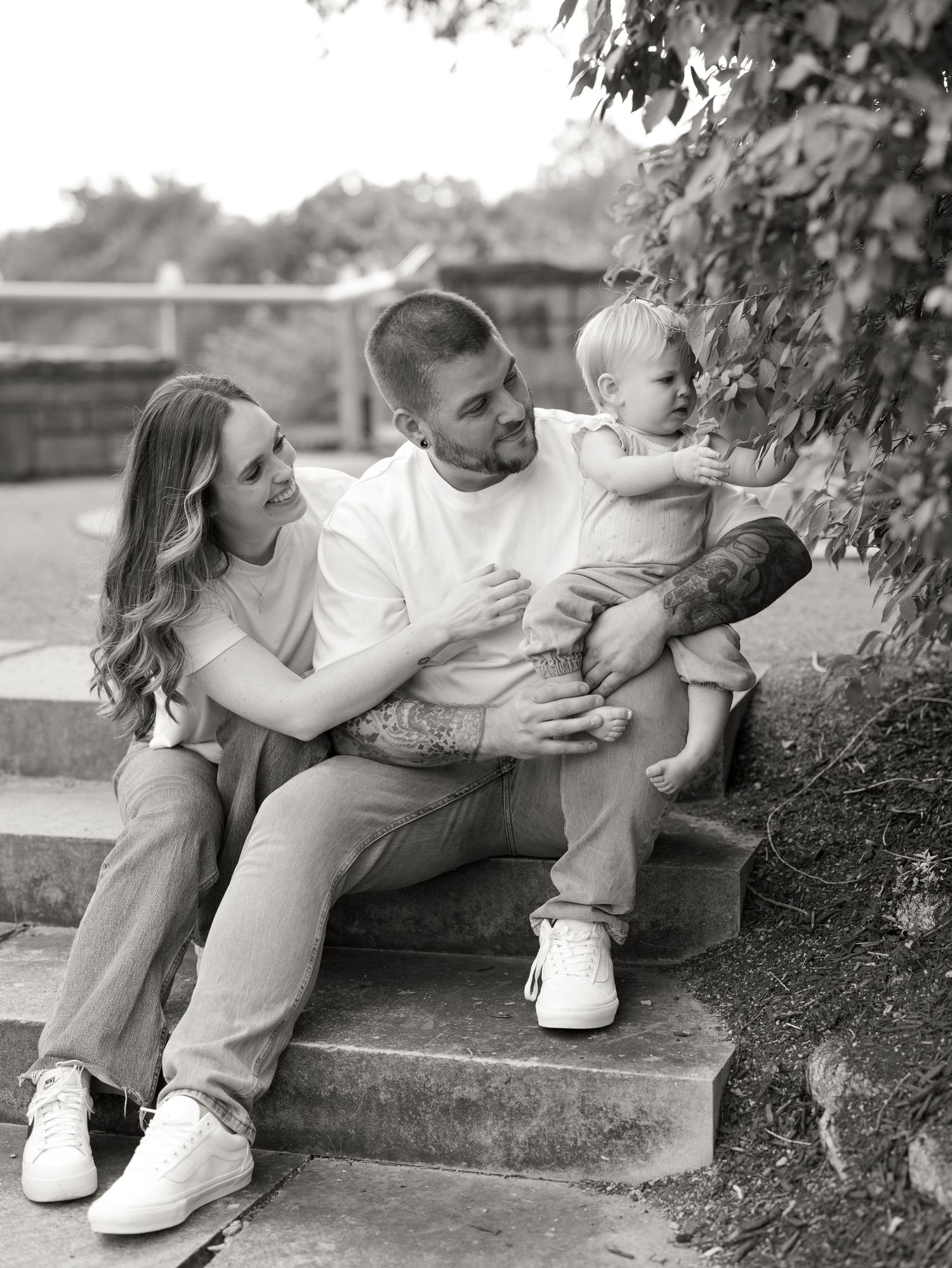 Black and white family portrait of couple sitting on stone steps with toddler reaching toward tree leaves.