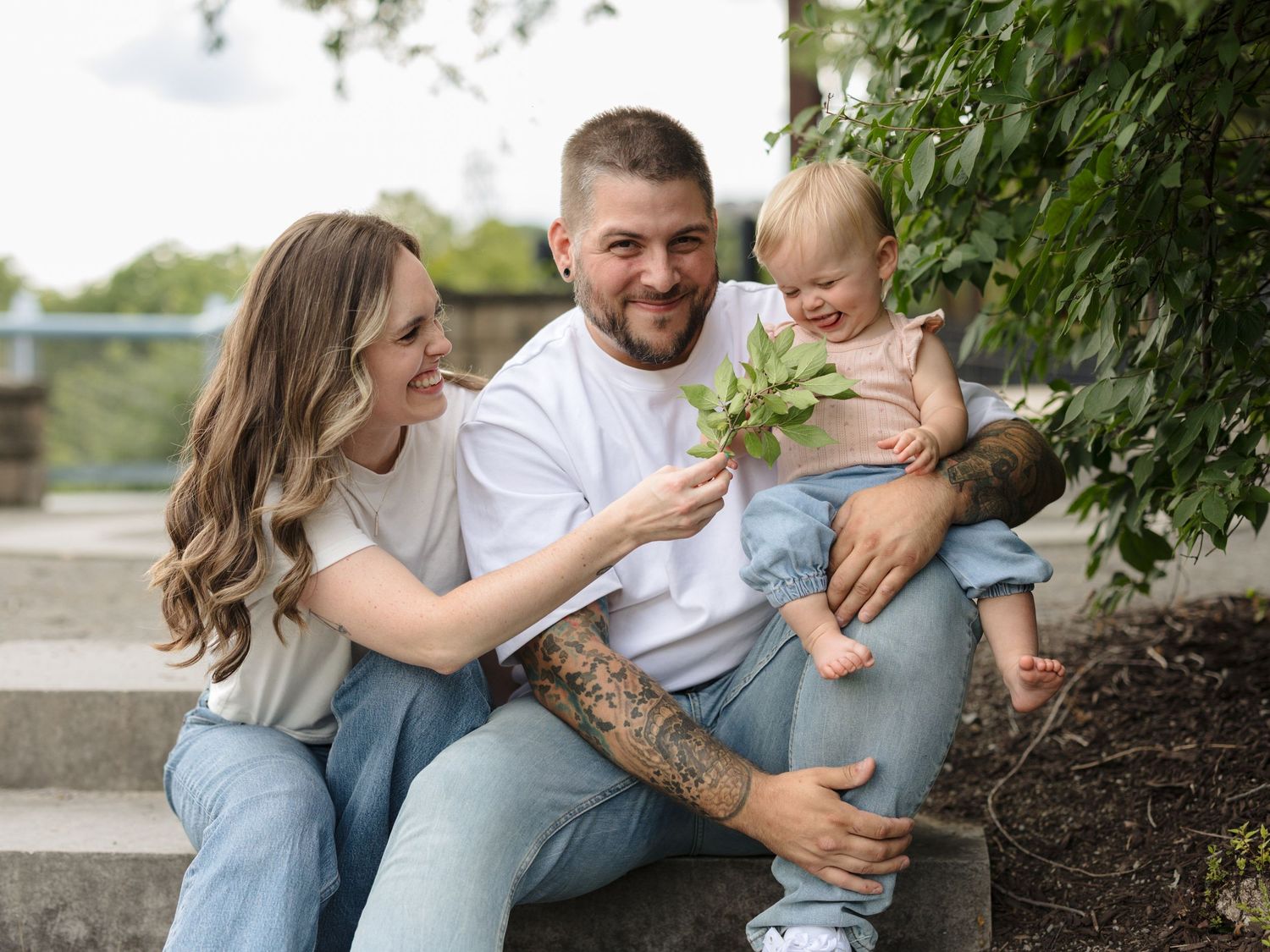 Happy young family with tattoed dad, smiling mom, and toddler sitting outdoors holding a green plant together.