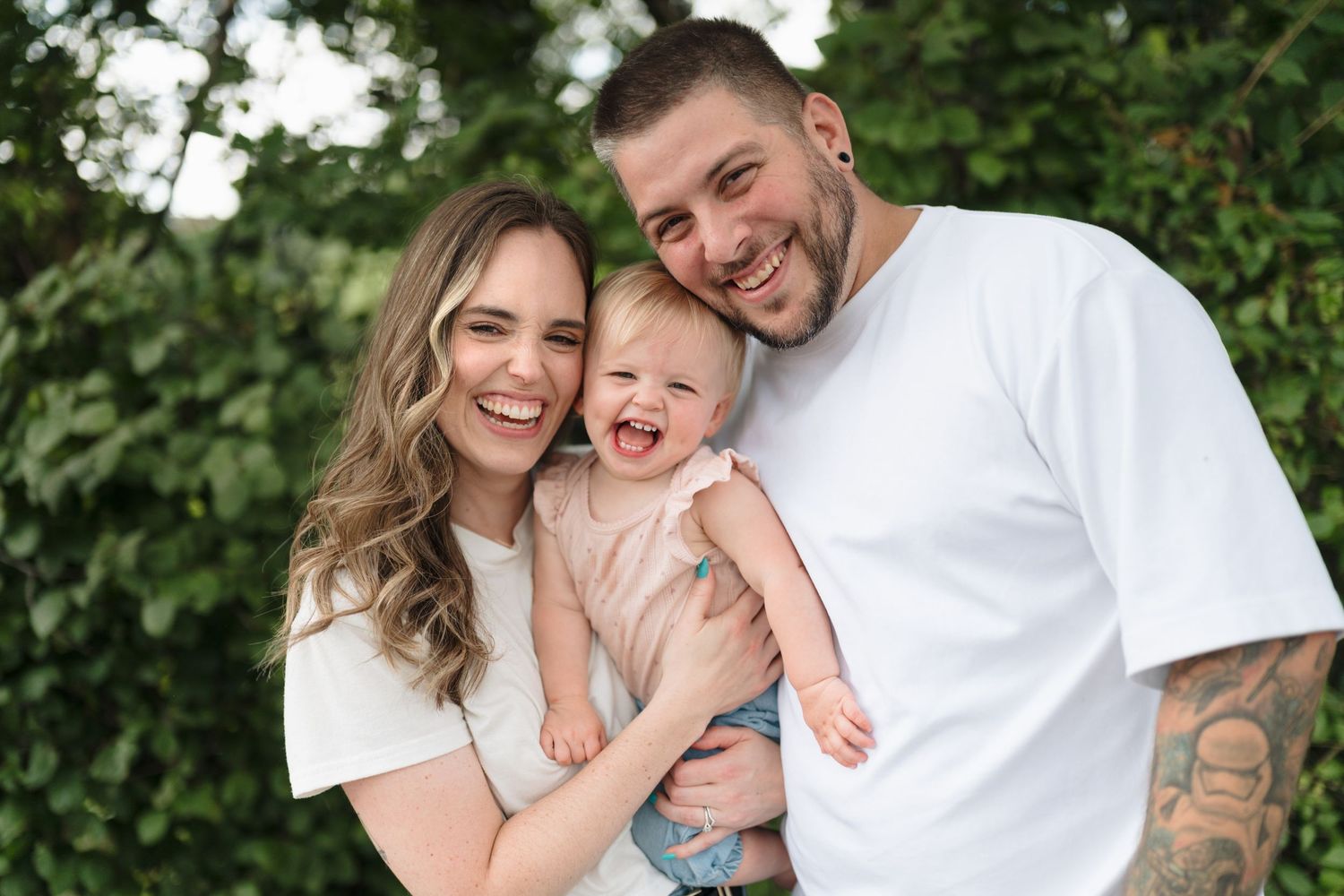 Happy young family with tattooed dad, smiling mom, and laughing baby posing outdoors with green foliage background.