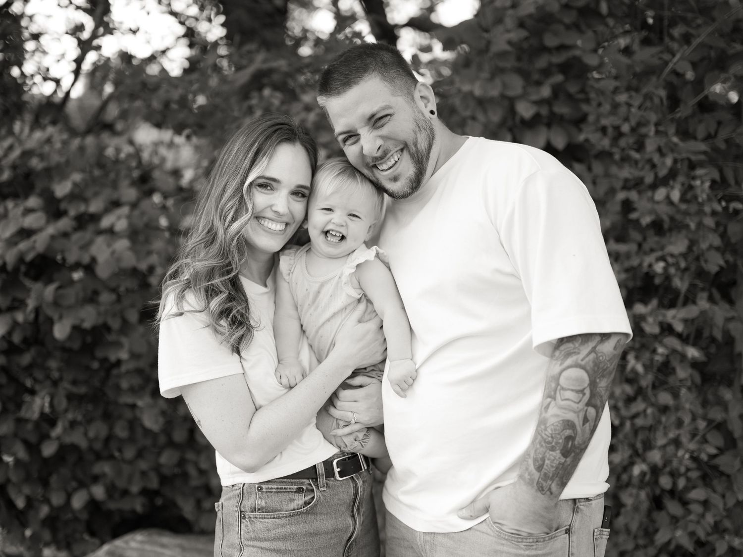 Black and white family portrait of smiling couple holding happy baby outdoors with lush green foliage background.