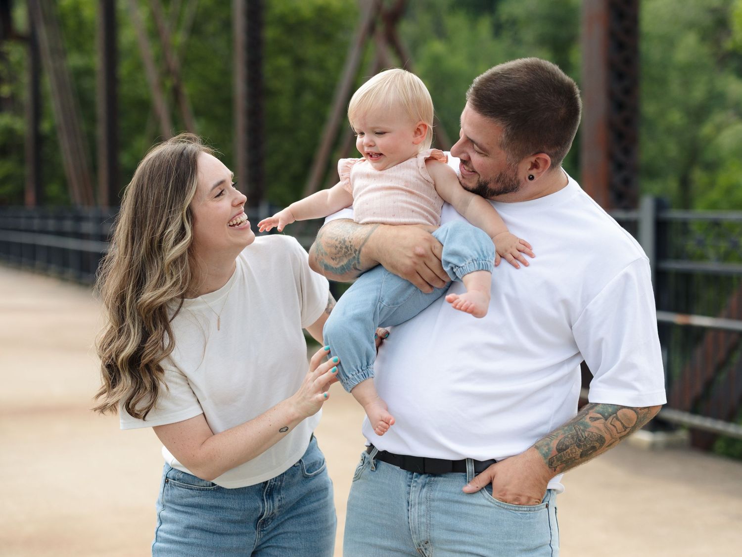 Happy young family with tattooed dad holding toddler baby while mom smiles outdoors near bridge.