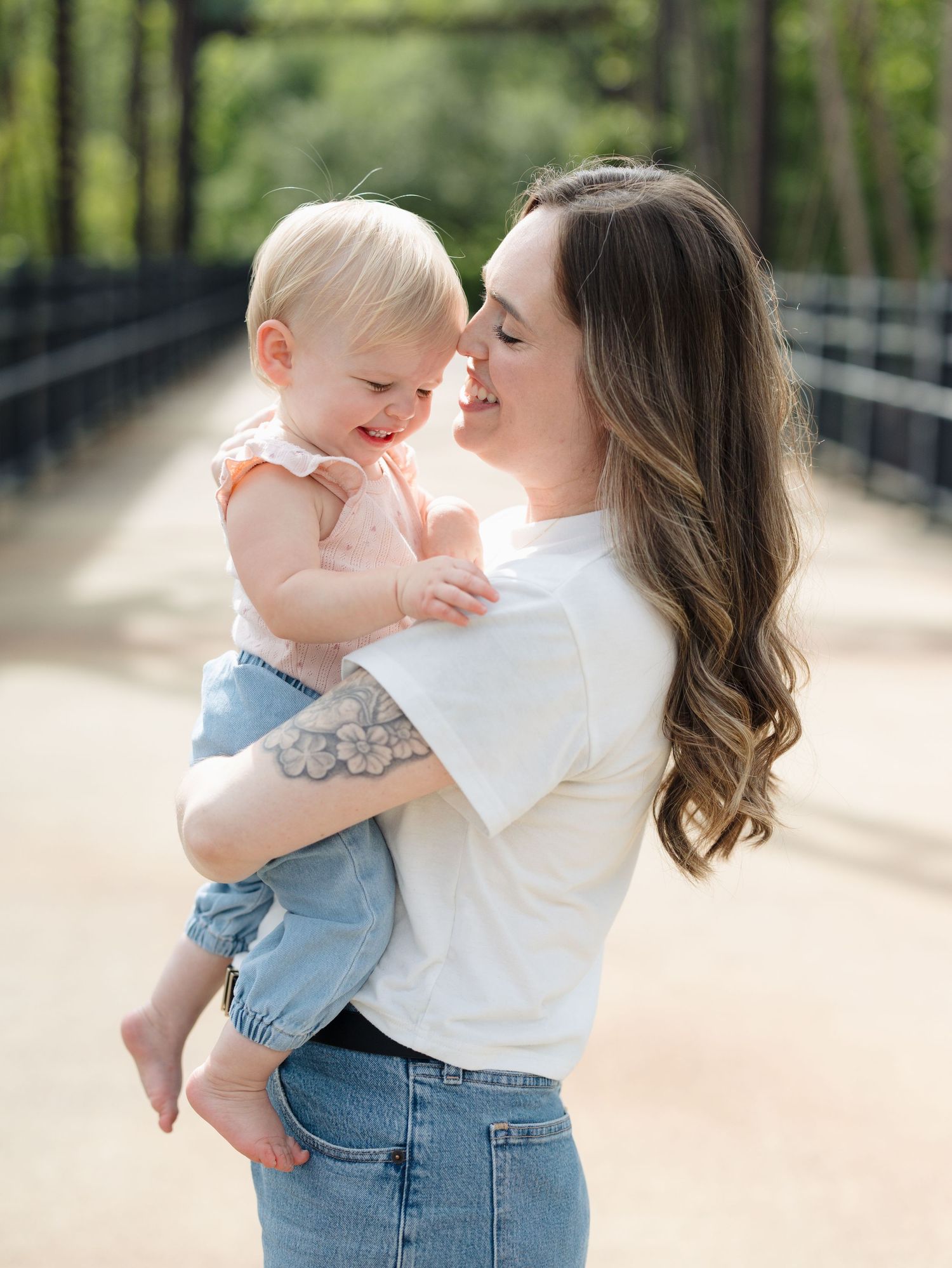 A young mother with long brunette hair holds her smiling baby girl on a sunlit bridge outdoors.