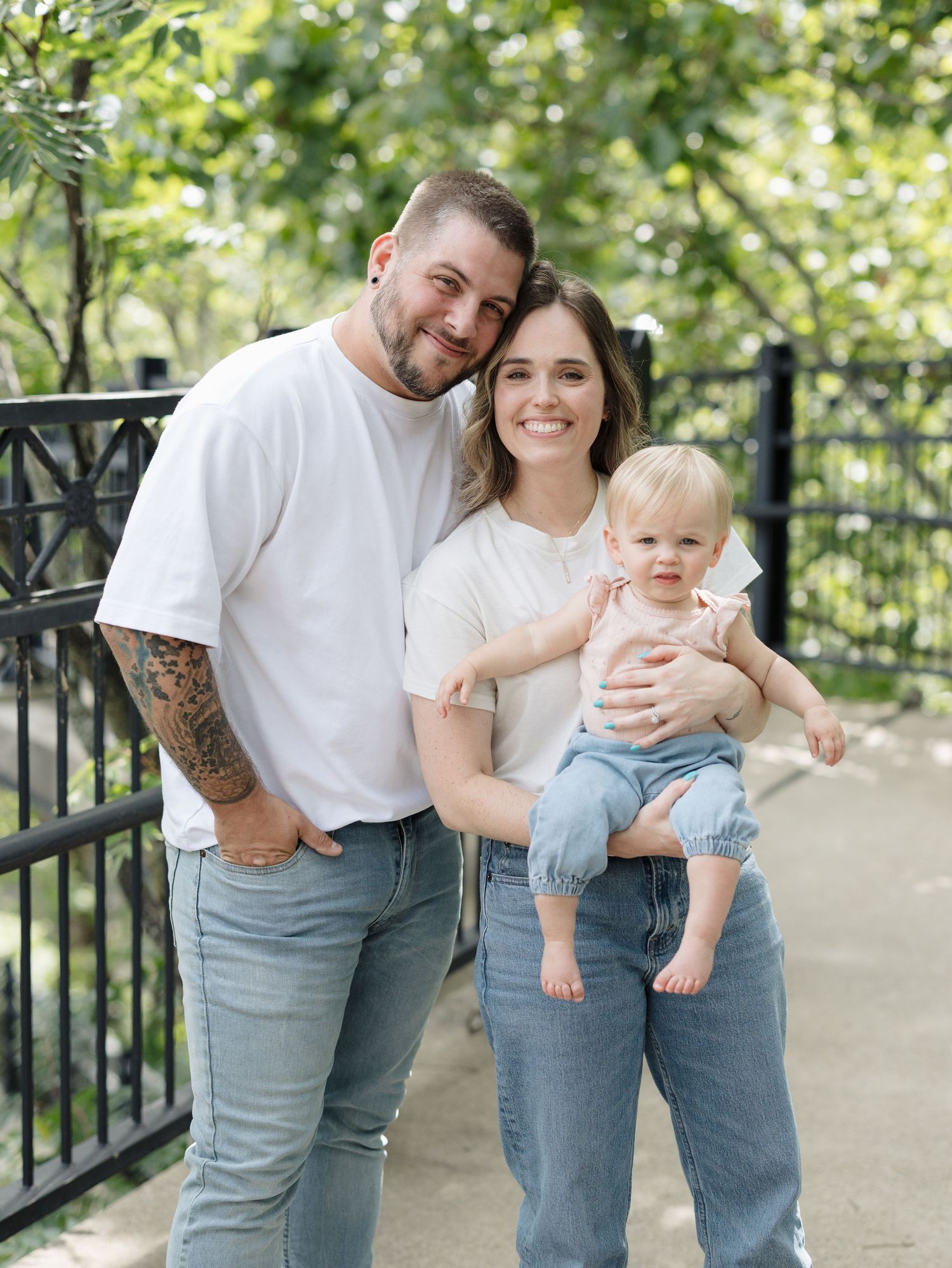 Young family with tattooed dad, mom, and baby girl posing outdoors in matching white tees and jeans near iron fence.