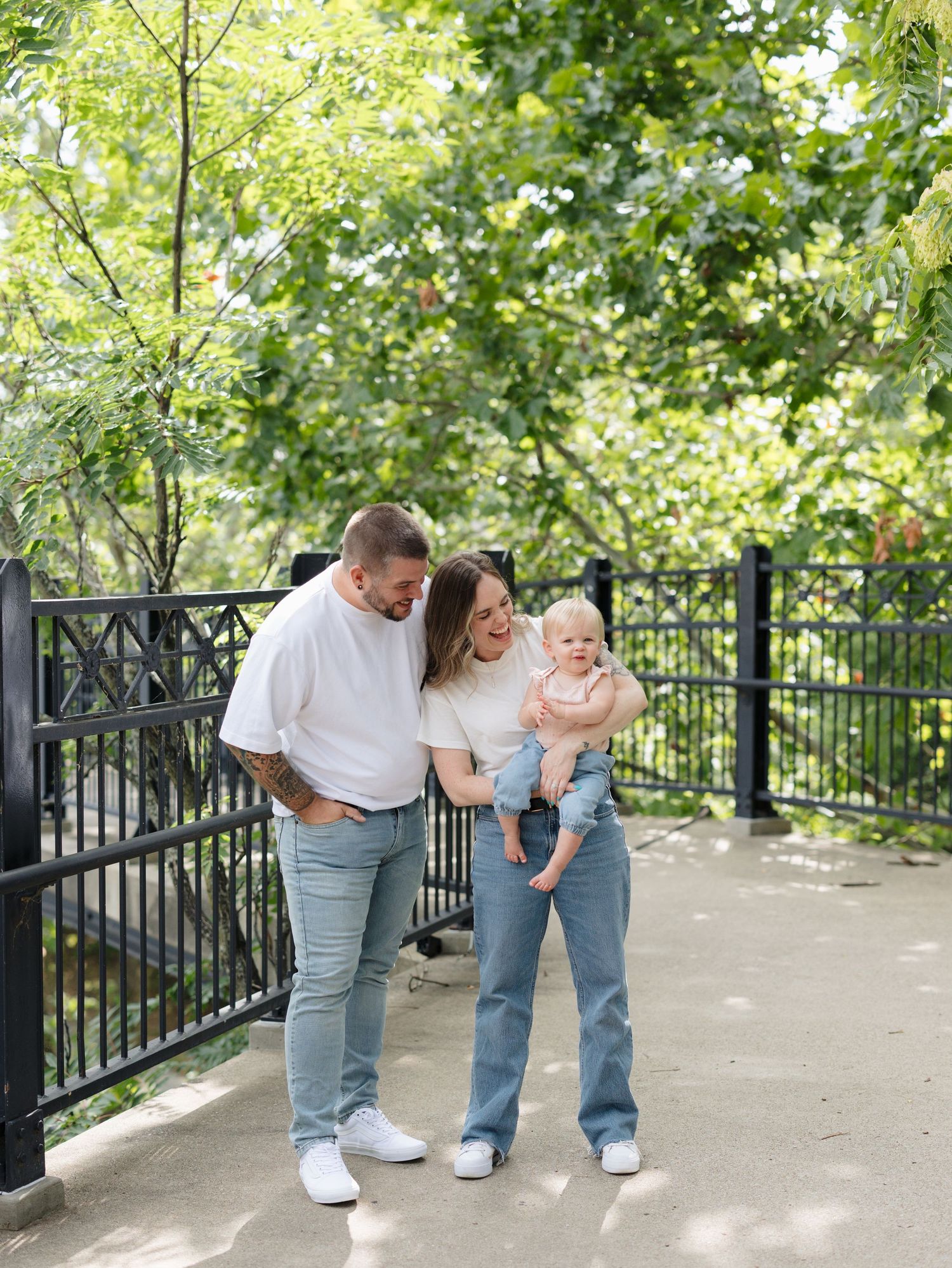 A young family with a baby poses on a park bridge surrounded by lush green trees on a sunny day.