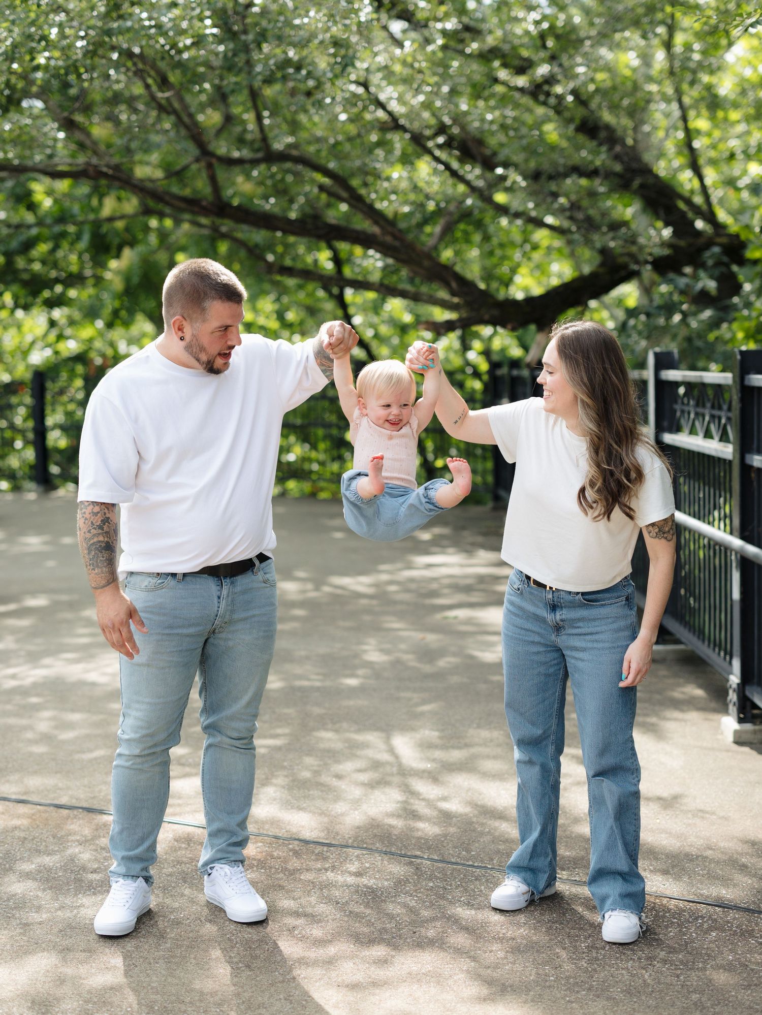 A young couple in matching white tees and jeans joyfully swing their baby between them on a sunny park bridge.