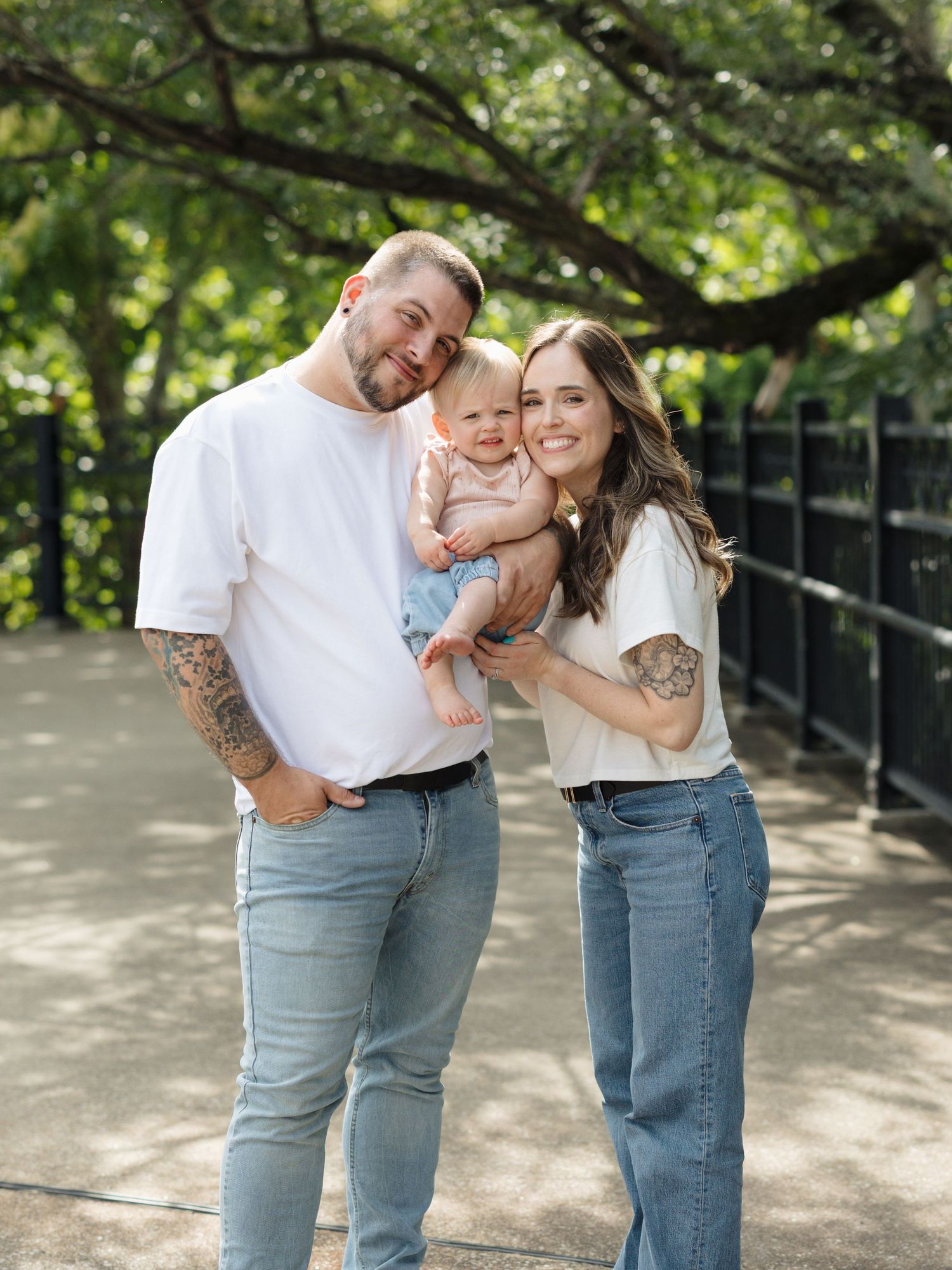 Smiling couple with sleeve tattoos holding adorable baby outdoors under green trees in coordinating white shirts and jeans.