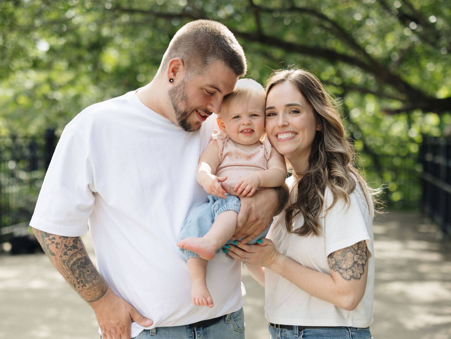 Happy young family with tattoos holding smiling baby outdoors in a lush green park setting.