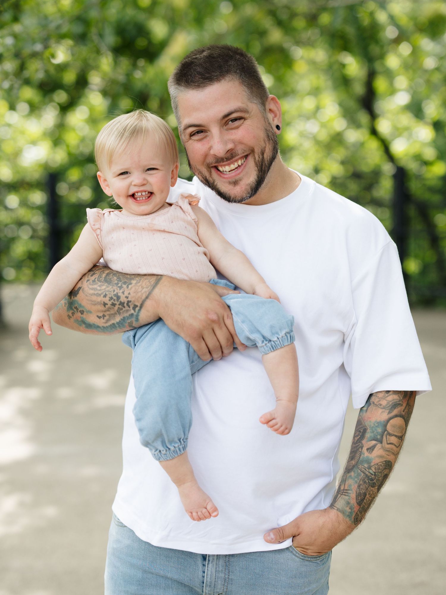 Smiling tattooed father holding happy baby girl outdoors in a park, wearing white t-shirt and jeans.