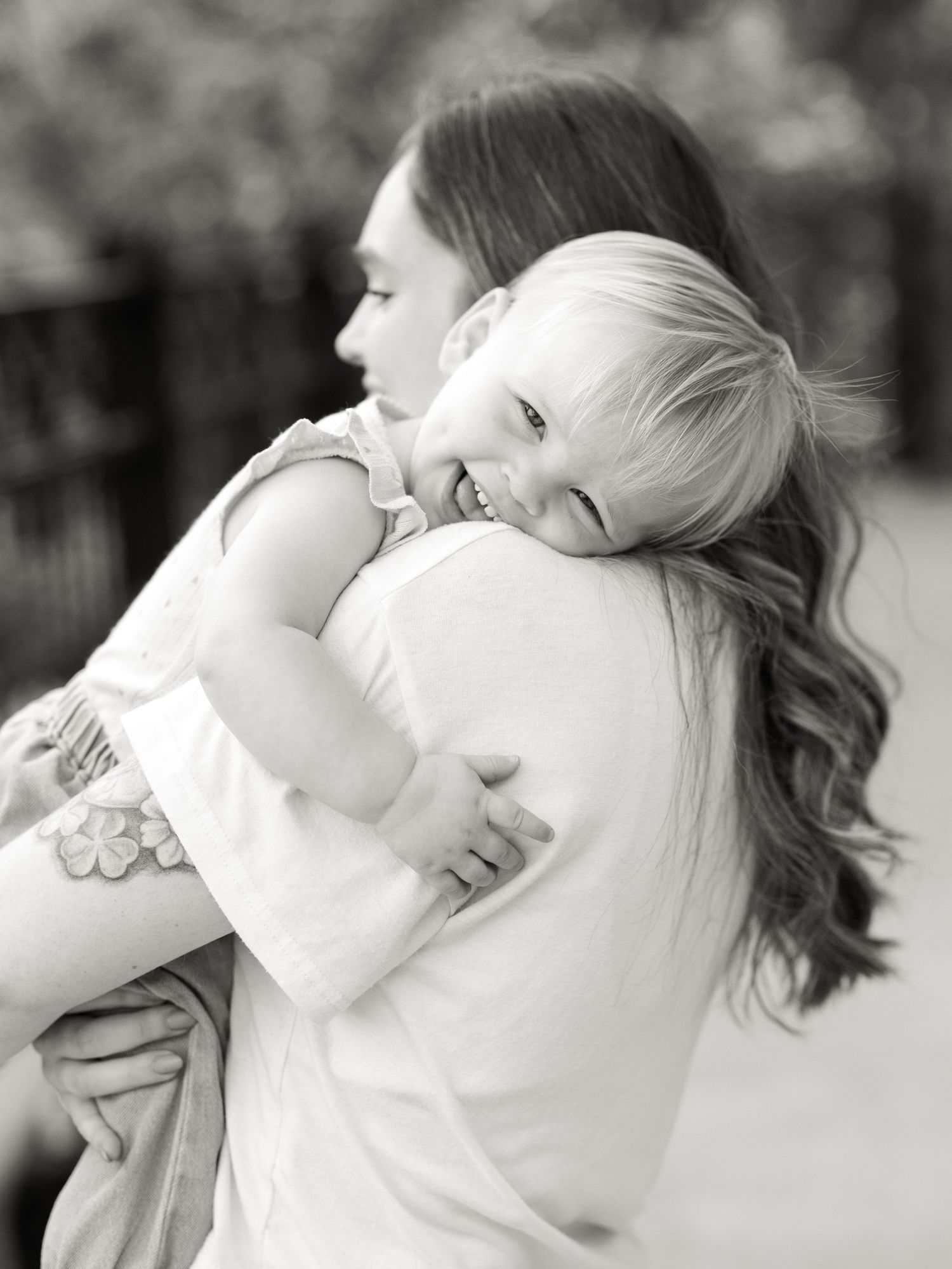 Black and white photo of a smiling toddler hugging a woman with long wavy hair outdoors.