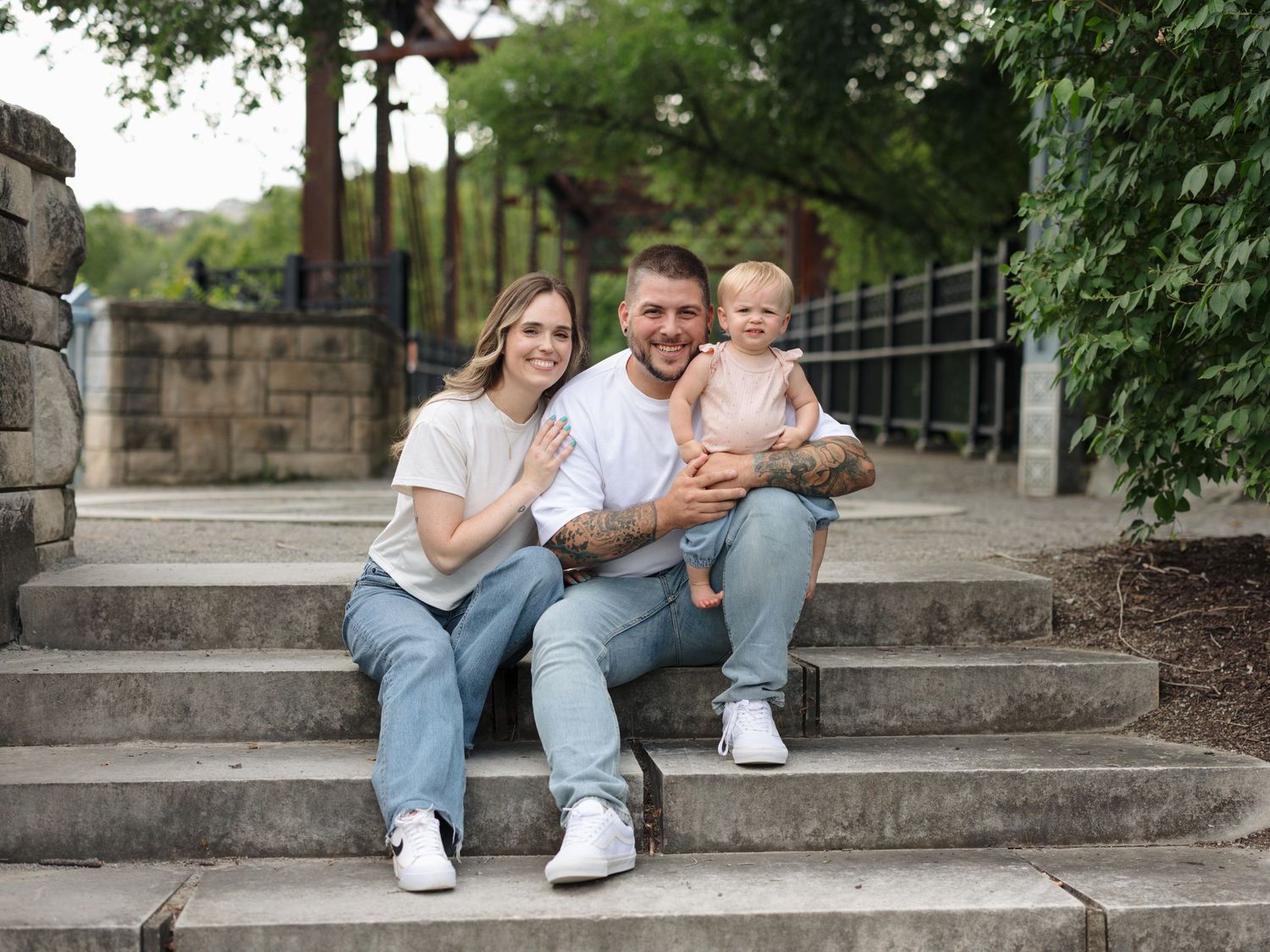 Young couple sitting on stone steps outdoors holding a baby, all wearing white shirts and jeans, smiling at camera.
