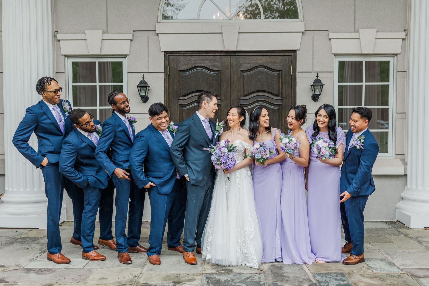 Wedding party poses outdoors, groomsmen in navy suits and bridesmaids in lavender gowns with bride in white.