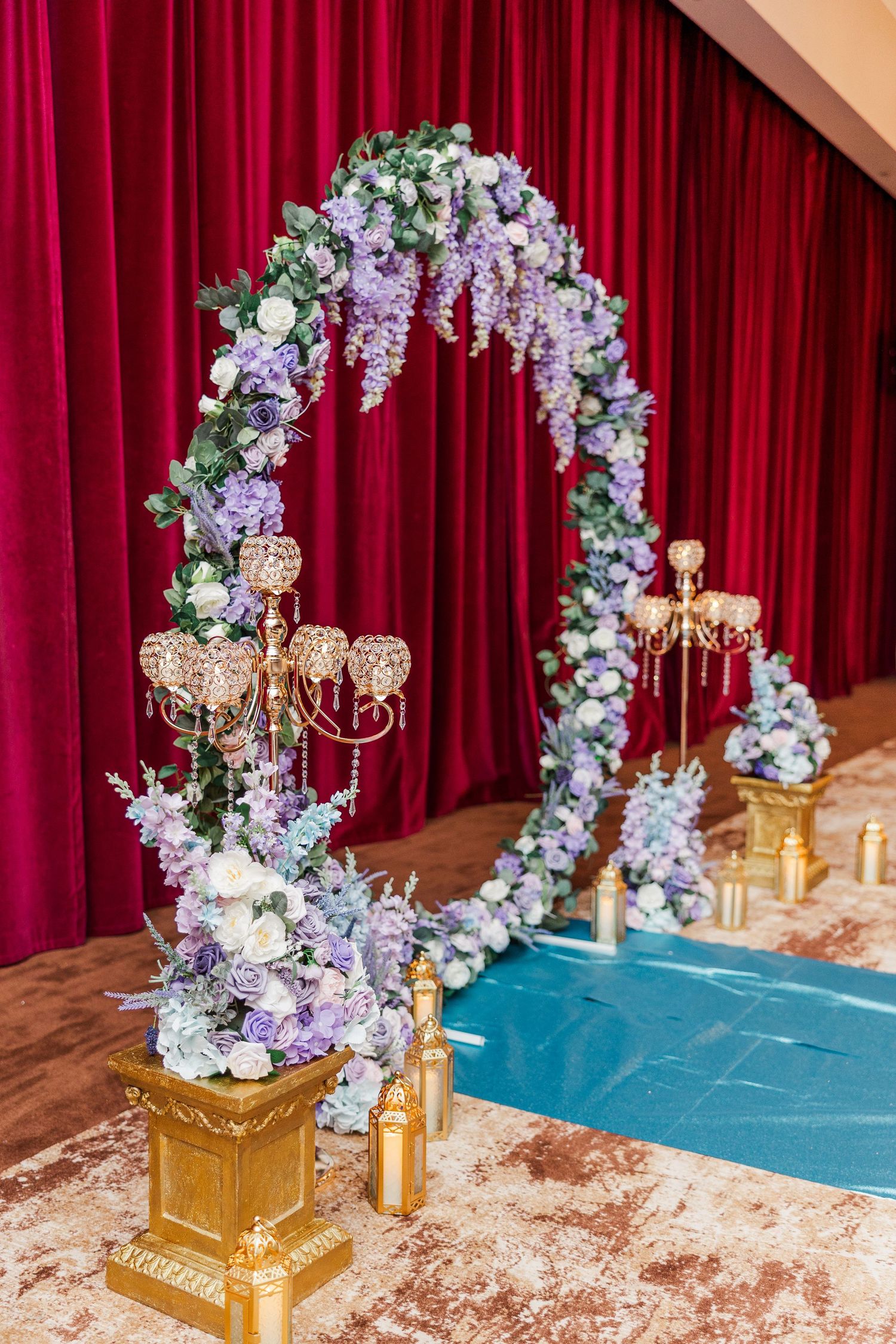 Elegant wedding arch adorned with purple and white flowers, gold candelabras, and blue aisle runner against red velvet curtains.