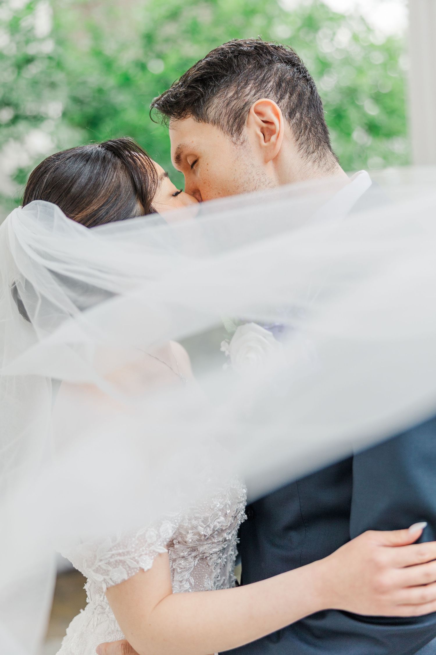 Bride and groom sharing a romantic kiss under flowing white veil on their wedding day.
