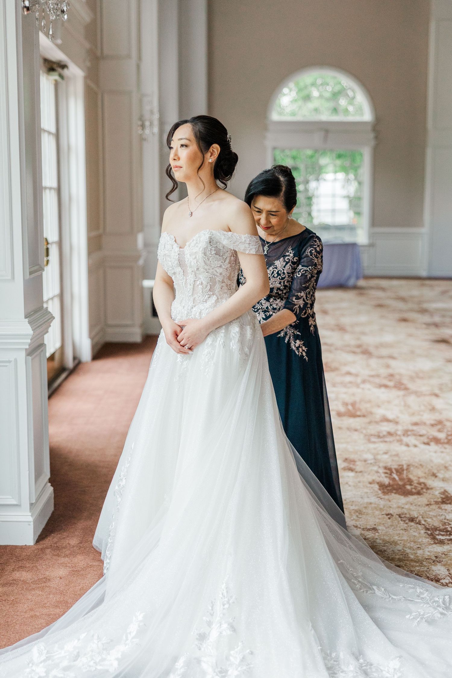 Bride in off-shoulder lace wedding gown being helped by woman in navy dress in elegant venue.
