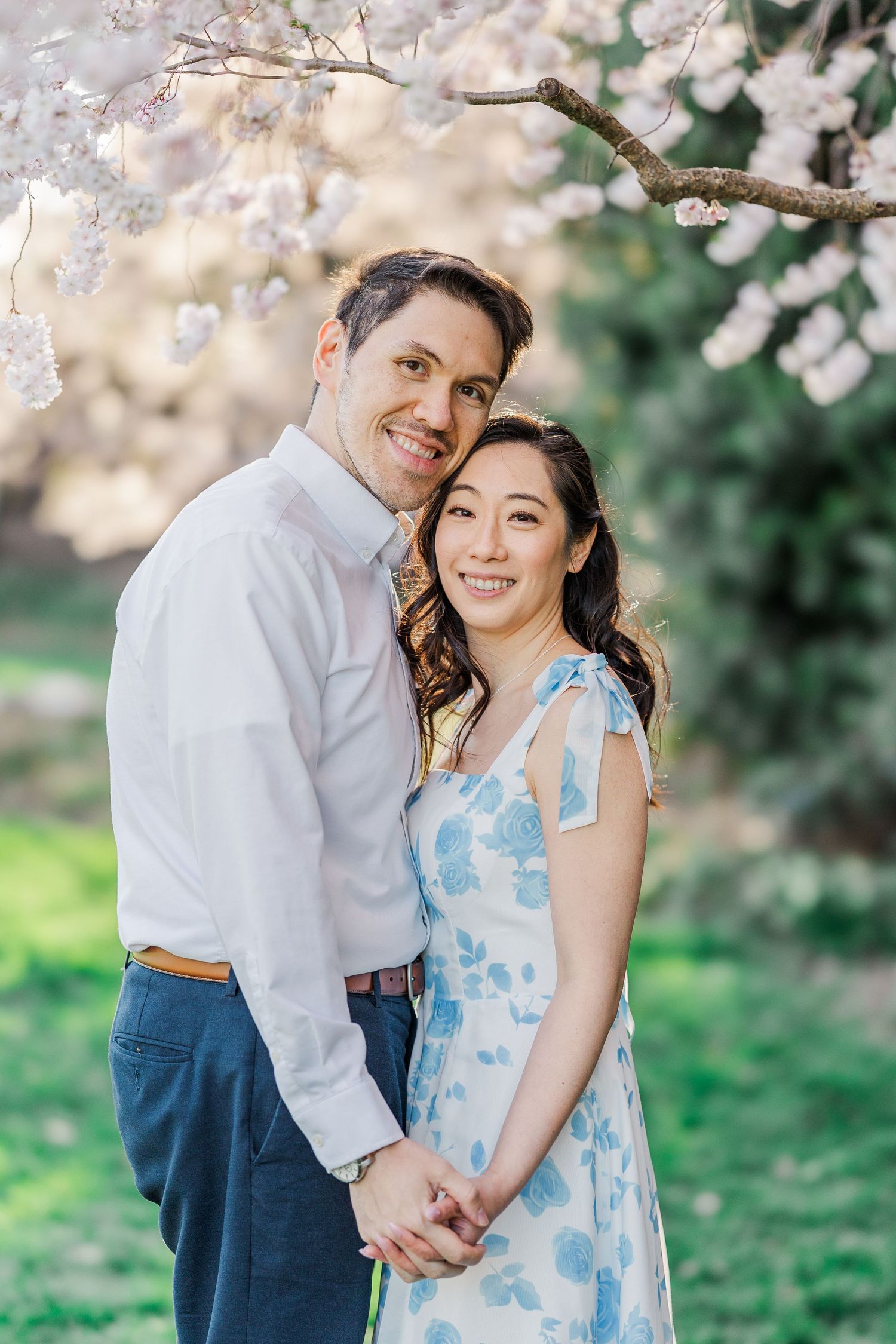 Couple embracing under cherry blossom tree, woman in floral dress and man in white shirt, smiling outdoors.