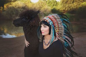 Woman in a turquoise and red feathered headdress poses beside a dark fluffy llama near a calm pond at golden hour.