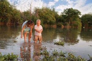 A woman and a toddler stand barefoot in shallow lake water, reflecting on the surface, with green trees and aquatic plants along the shore.