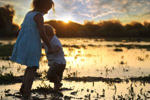 Two young children hold hands while wading through shallow, reflective water as the sun sets behind them.