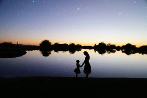 Silhouette of a mother holding her young daughter's hand beside a calm lake under a starry twilight sky.