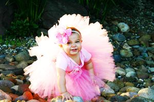 Smiling baby girl in a pink tutu skirt and flower headband sitting on smooth river rocks outdoors.