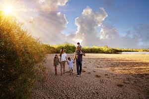 Family of five walking hand in hand on cracked dry earth at sunset, with a child riding on a parent's shoulders.