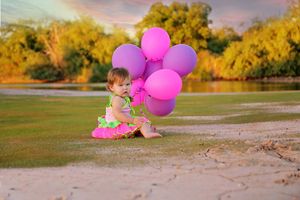 Baby girl in a pink tutu and green top sits on grass holding a bunch of pink balloons beside a river.