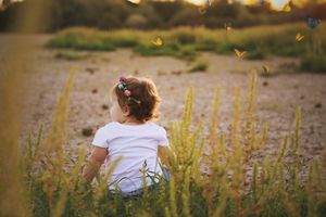Toddler with a flower hair clip sits in tall golden grass at sunset, watching butterflies in the field.