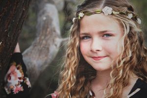 Smiling young girl with long curly blonde hair wears a delicate flower crown in a soft-focus outdoor portrait near a tree.