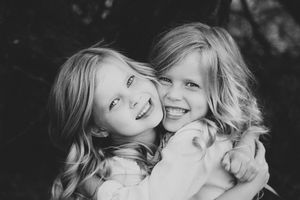 Two smiling young girls with long wavy hair hugging each other in a joyful black and white portrait.