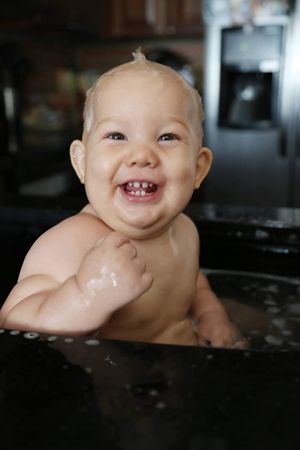 Smiling baby with wet hair and a toothy grin sits in a sink-style baby bath filled with water in a cozy kitchen.