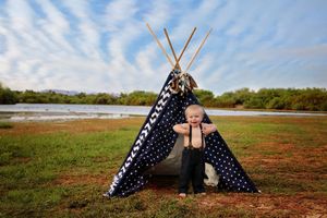 Smiling toddler boy in suspenders stands in front of a navy polka dot teepee on a grassy lakeside field.
