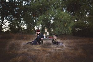 Two people in dark clothing sit beside a small table with lit candles in a dry grass clearing surrounded by trees at dusk.