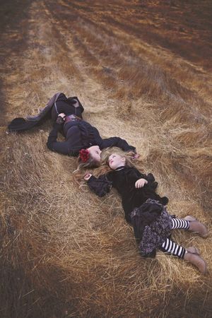 Two girls in dark vintage clothing and striped tights lie head to head on dry golden straw in an autumn field.