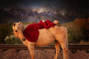Woman in a red gown reclines on a palomino horse beside railroad tracks under a starry Milky Way night sky.