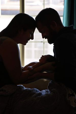 Silhouette of parents touching foreheads while gently holding their newborn baby beside a bright window.
