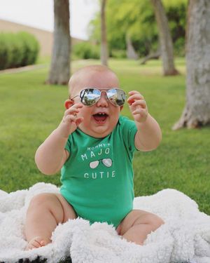Smiling baby in mirrored aviator sunglasses and a green Mommy's Major Cutie bodysuit sitting on a white fuzzy blanket on a sunny park lawn.