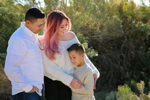 A man, a woman with pink-dyed hair, and a young boy share a warm family embrace outdoors in front of green shrubs in soft natural light.