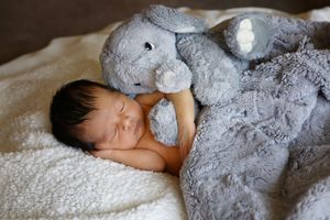 Sleeping newborn baby hugging a soft gray stuffed elephant on a cozy white blanket.