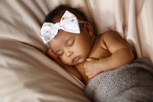 Sleeping baby girl wearing a white floral bow headband, resting peacefully on a soft beige blanket with a gray knit wrap.