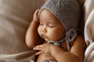 Sleeping baby wearing a soft gray knitted bonnet with ties, resting peacefully with one hand raised near the face on a neutral blanket.