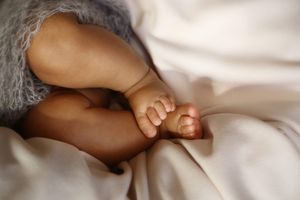 Close-up of a newborn baby's tiny bare feet and legs resting on soft white linens beside a gray knit garment.