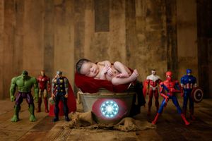 Newborn baby sleeping on a red cape in a wooden crate with a glowing Iron Man arc reactor, surrounded by superhero action figures.