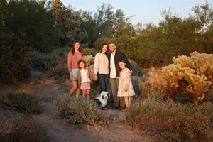 A family of four with a small fluffy dog poses on a desert trail surrounded by green shrubs and cacti in warm golden evening light.
